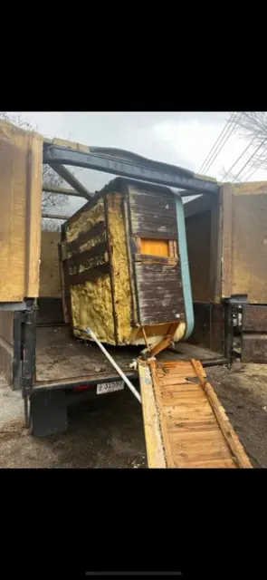 A dilapidated, partially dismantled wooden shed structure sits inside the back of a truck, connected to a wooden ramp.