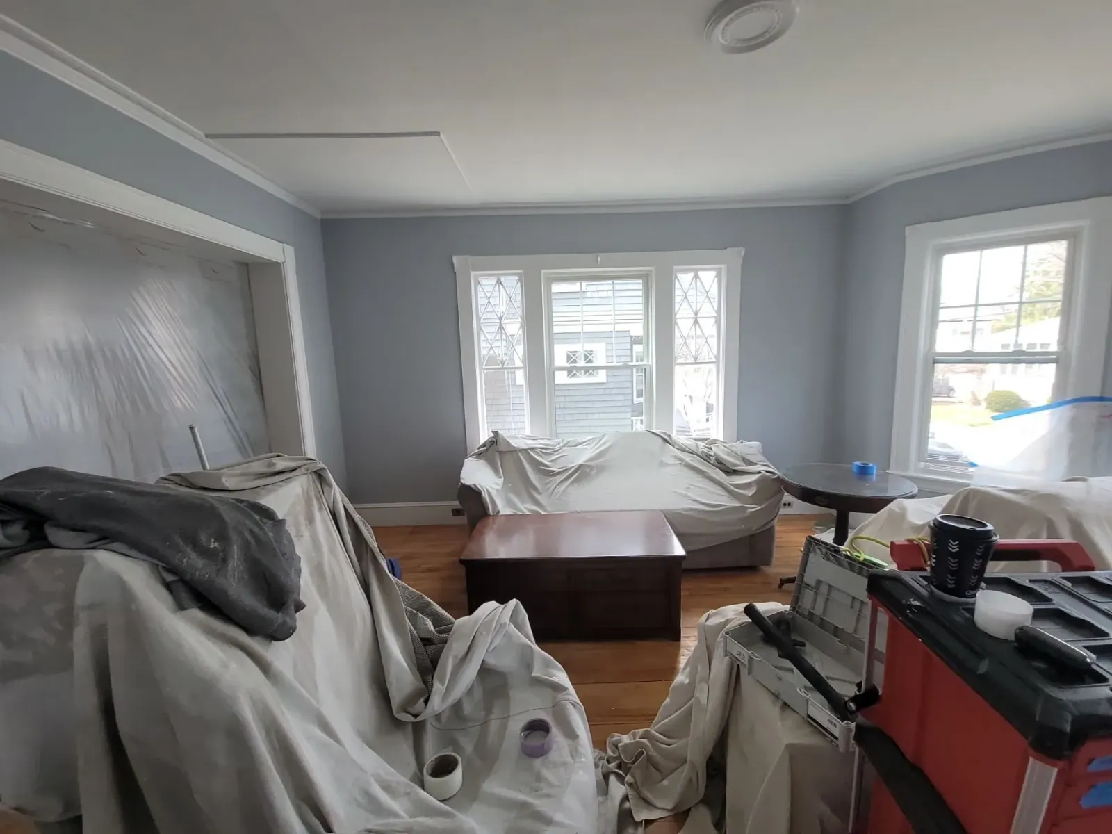 Gray-walled living room with covered furniture, a coffee table, and large windows letting in daylight