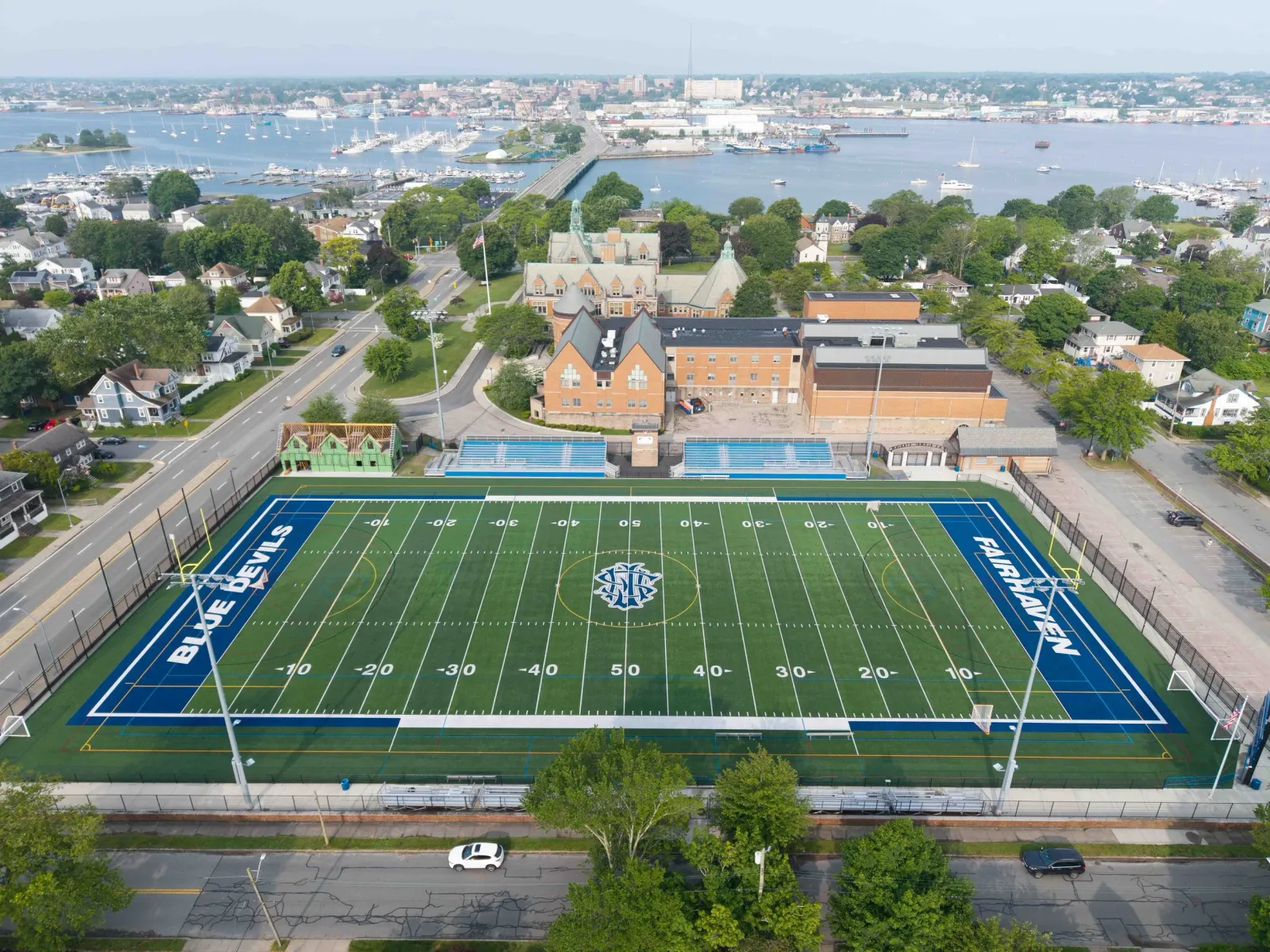 Aerial view of a football field with the words 'BLUE DEVILS' and 'FAIRHAVEN' on the sidelines, surrounded by trees, residential houses, and a harbor with boats in the background.