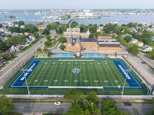 Aerial view of a football field with the words 'BLUE DEVILS' and 'FAIRHAVEN' on the sidelines, surrounded by trees, residential houses, and a harbor with boats in the background.