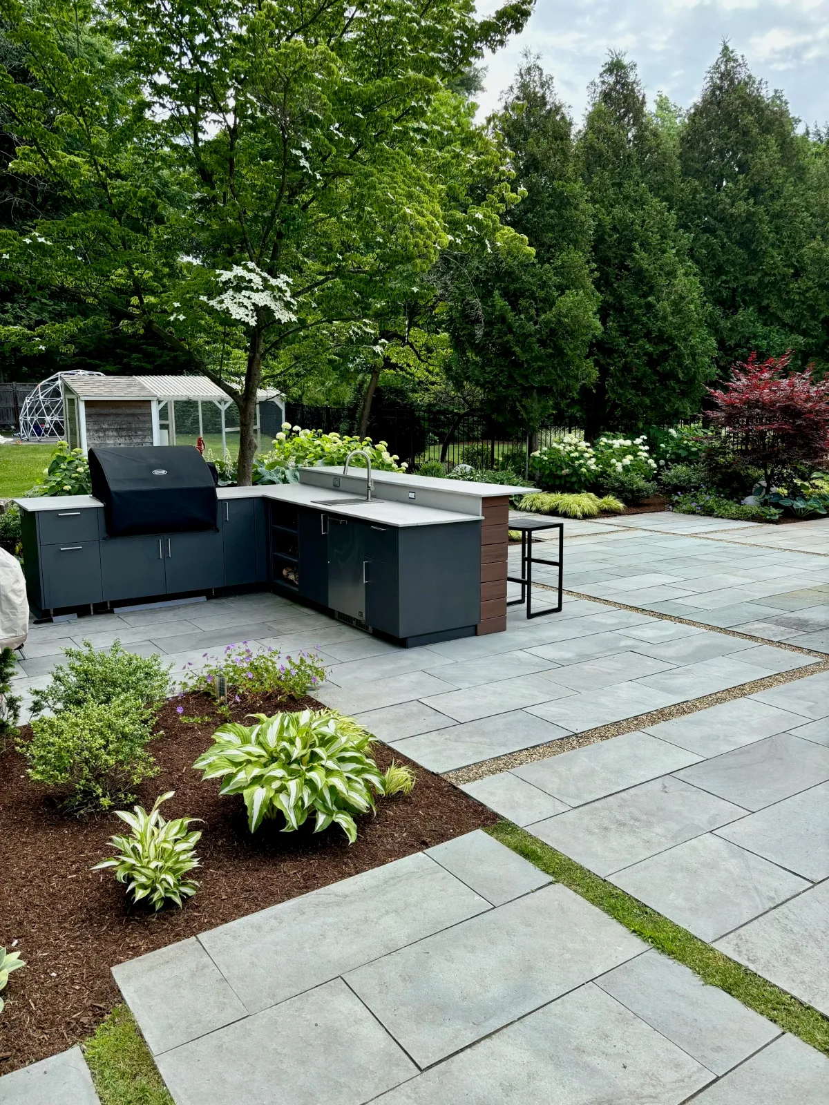 An modern outdoor kitchen with grill station, sink. fridge and cabinet and counterspace along with bar seating at this Andover landscape transformation combine with blue mist granite pavers and thoughtful landscape design to create a series of patio