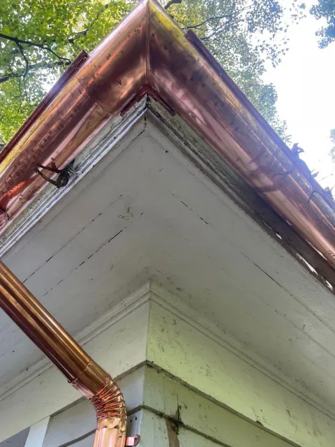 Copper gutter and downspout on a white house corner under trees, showing rust and weathering