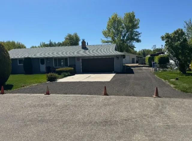 A gray house with a driveway, grass, and trees on a sunny day. Orange cones line the driveway.