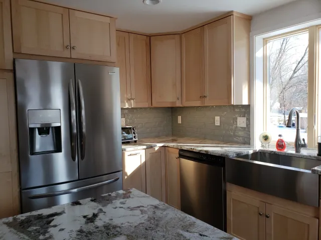 Kitchen with light wood cabinets, stainless steel refrigerator, granite countertop, gray subway tile backsplash, and a large window above a farmhouse sink.