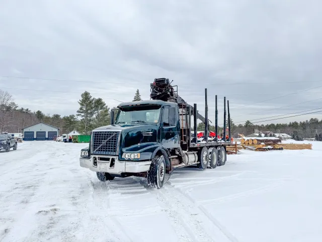 A black logging truck with a crane on top parked on snow-covered ground in a rural area with trees and buildings in the background. Tree Service Maine, Learned Tree Service