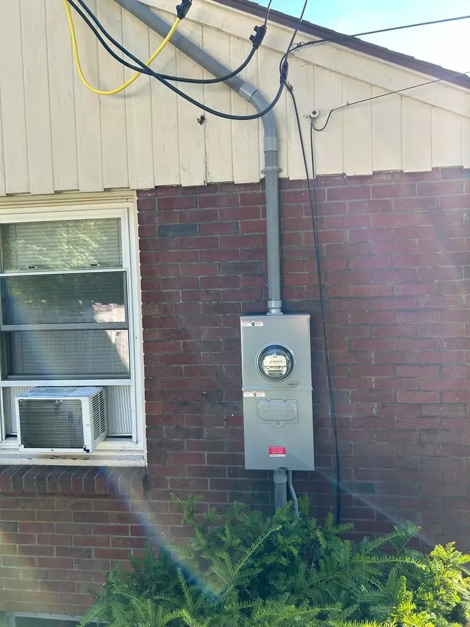 Electric meter and utility box on red brick wall with window and air conditioning unit nearby.