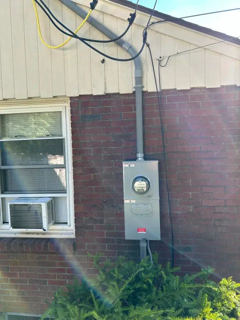 Electric meter and utility box on red brick wall with window and air conditioning unit nearby.