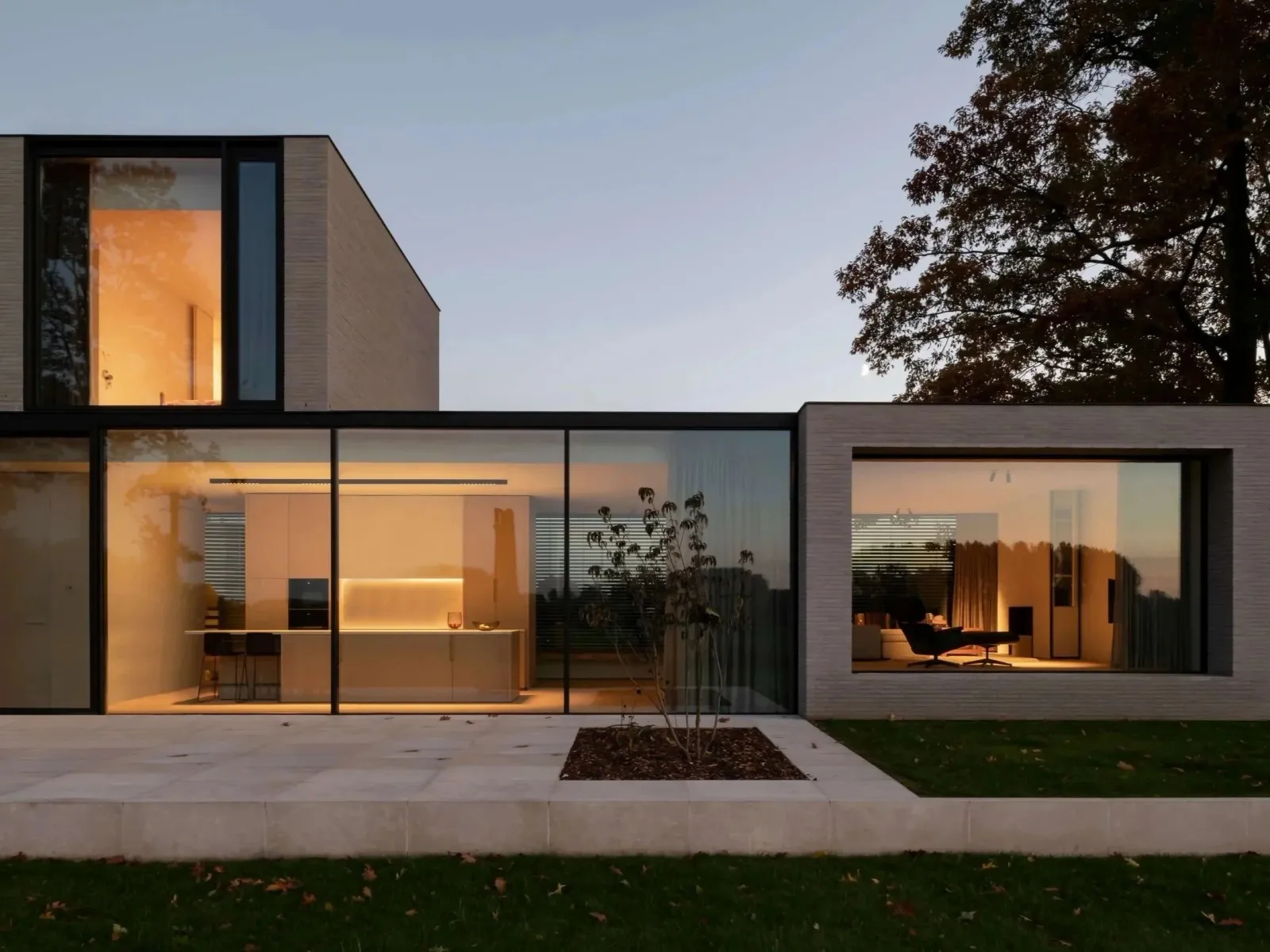 Modern house with large glass windows and interior lighting seen at dusk, with a patio, small tree, and trees in the background.