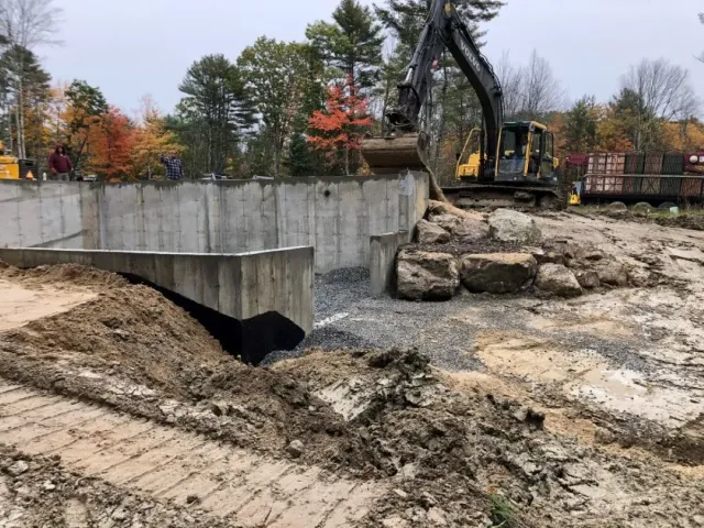 A large excavator is digging a hole in the ground in front of a concrete wall.