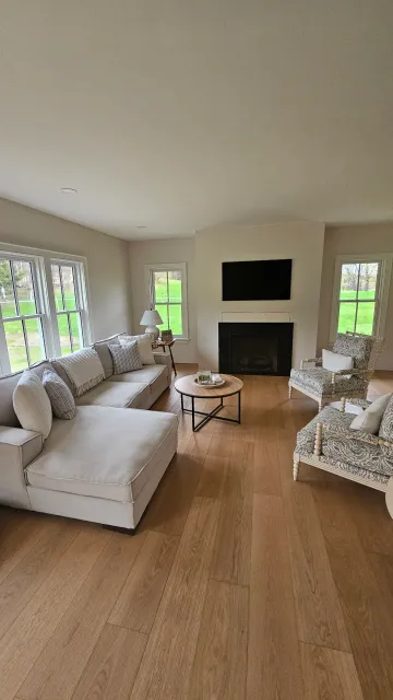 Living room with cream sofa, patterned armchairs, wood flooring, fireplace, mounted TV, windows with greenery outside, and a small round coffee table.