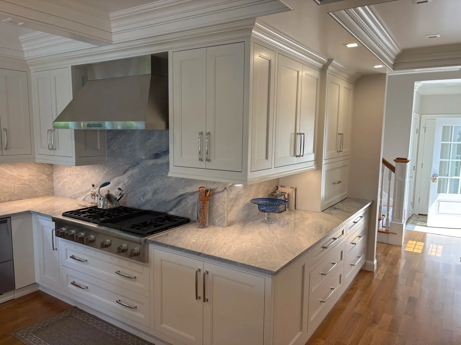 Modern kitchen with white cabinetry, marble countertops, stainless steel appliances, hardwood floors, and a view of a hallway with a staircase.