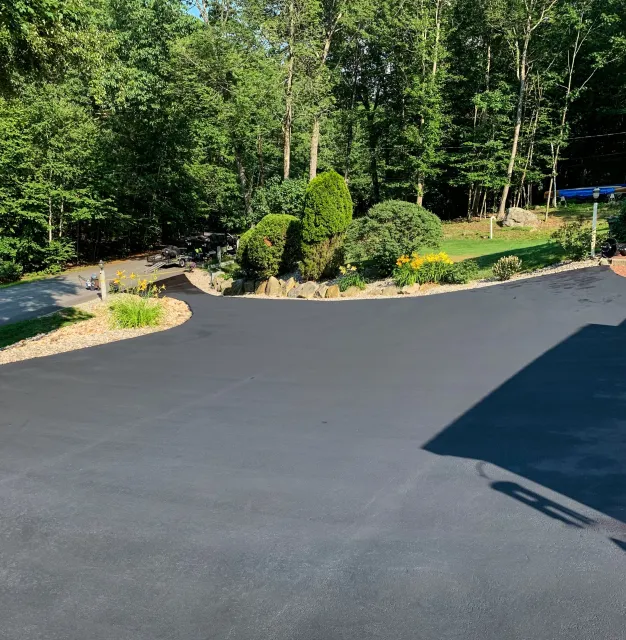Freshly paved dark asphalt driveway with landscaped flower beds and bushes, surrounded by green trees and a forest in the background.