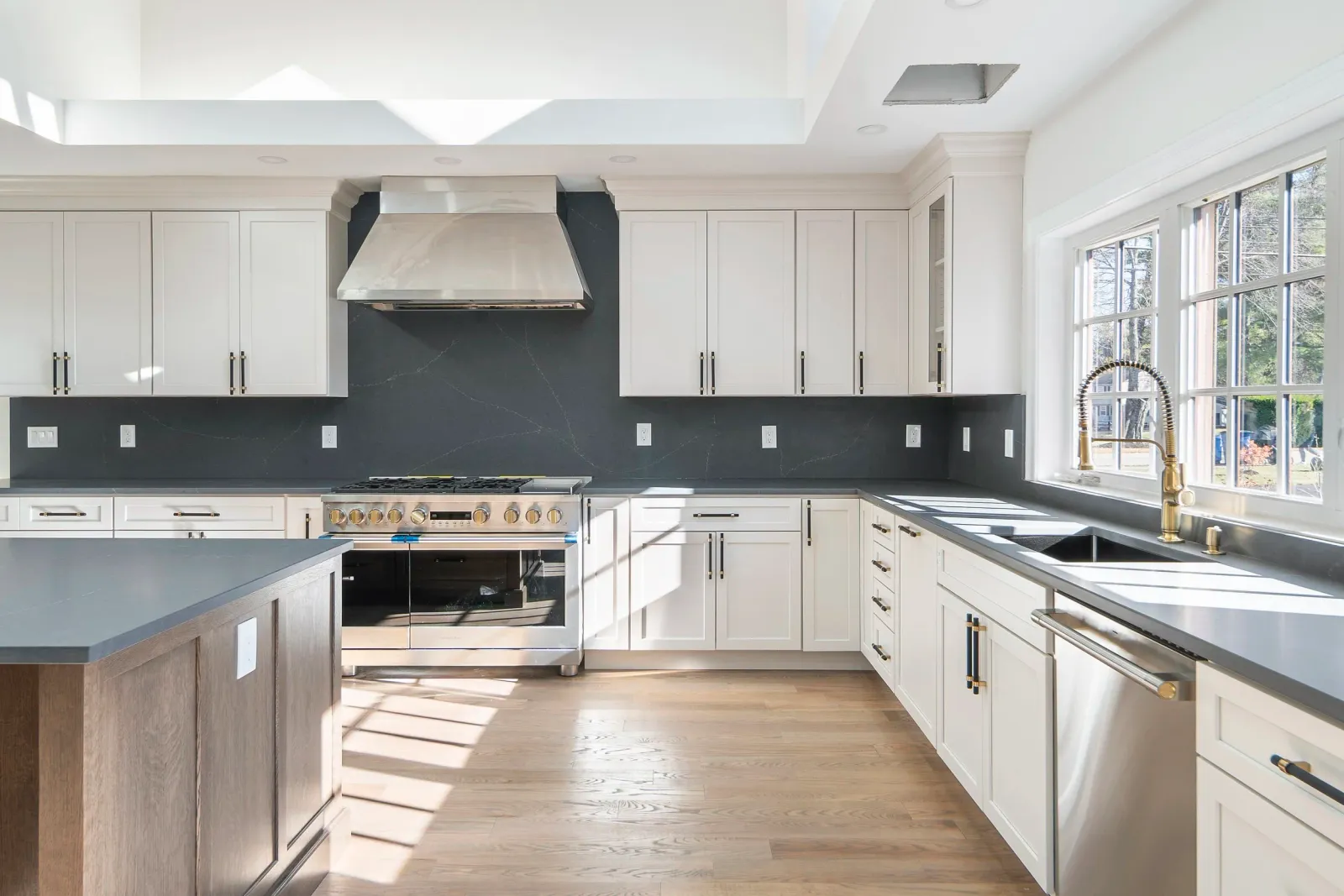 Modern kitchen with white cabinets, black countertops, large window, stainless steel appliances, and wood flooring.