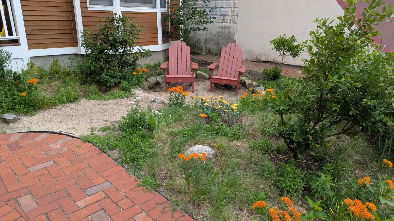 Two red Adirondack chairs on a small dirt patch in a garden surrounded by green plants and colorful flowers, next to a brick pathway.