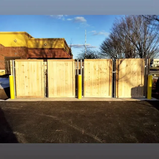 Wooden dumpster enclosure with yellow poles, in front of a building with blue sky.