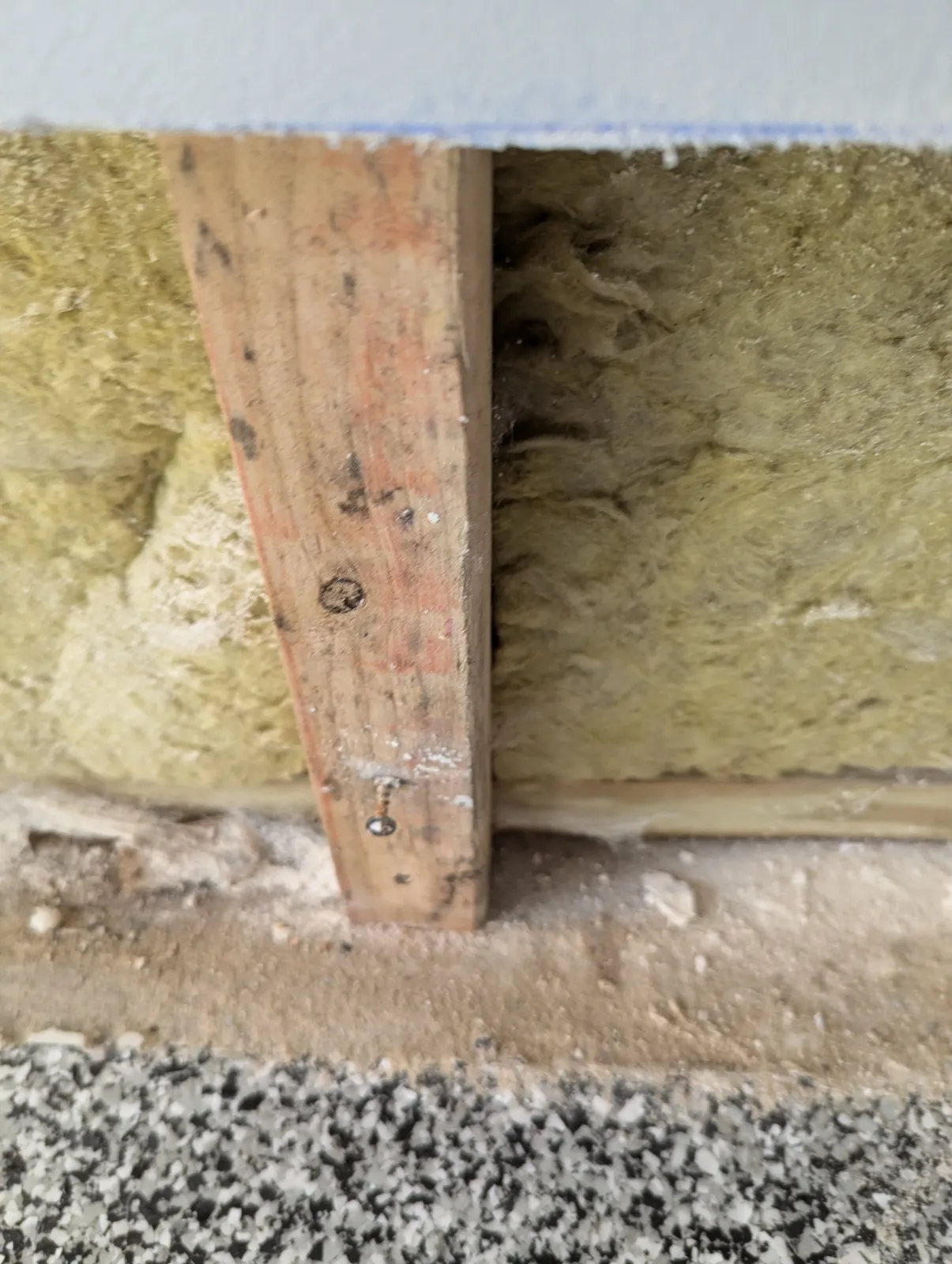 Close-up view of a corner of a room showing a wooden level tool placed against the wall, with yellow insulation and drywall on either side, and some debris and gravel on the floor.