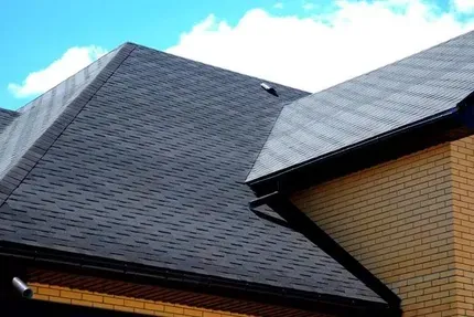 Dark asphalt shingle roof on a house, with black gutters and tan brick wall.