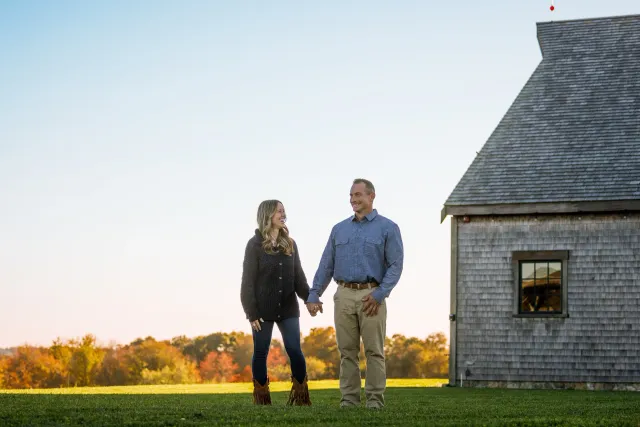 A couple holding hands outside near a barn during sunset, smiling at each other.