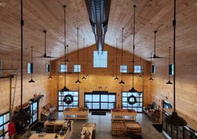 A wide-angle view of a wooden interior with high ceilings, multiple windows, and overhead lighting. The space features counters and produce displays, with wreaths hanging on the doors.