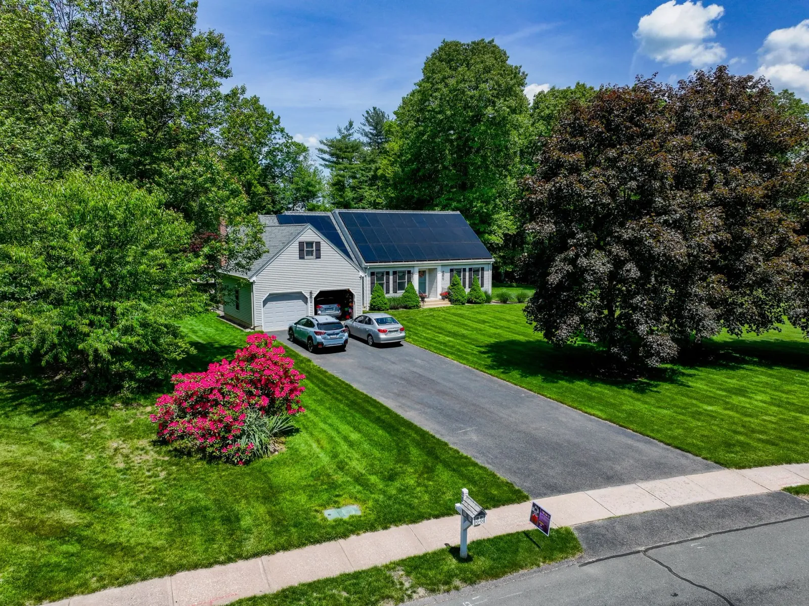 Suburban house with a driveway, parked cars, and lush green yard with trees and pink flowers