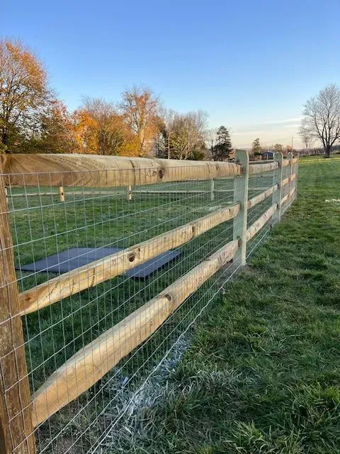 Wooden post-and-wire fence bordering a grassy field at sunset, with autumn trees in the background