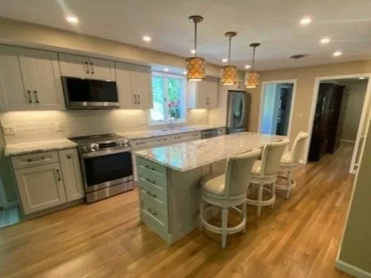 Modern kitchen with light wood cabinets, stainless steel appliances, large island with granite countertop, and barstool seating. Hardwood floors and pendant lighting enhance the space.