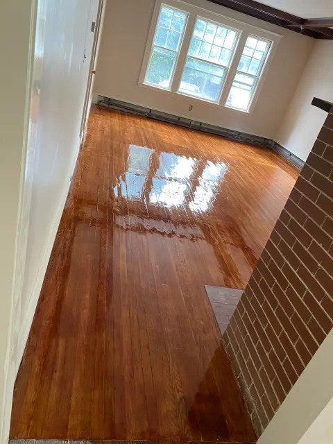 A room with polished, light-brown wooden floors reflecting the windows, adjacent to a brick wall and cream-colored walls.