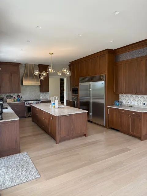 A spacious modern kitchen with wooden cabinets, a large central island, stainless steel refrigerator, and hardwood flooring. There is a stylish chandelier hanging from the ceiling.